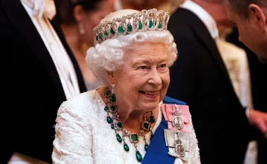 A smiling elderly woman wearing a white dress, emerald jewelry, and a tiara at a formal event.