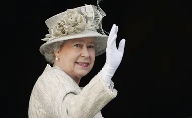 An elderly woman in a floral hat and white gloves waves gracefully.