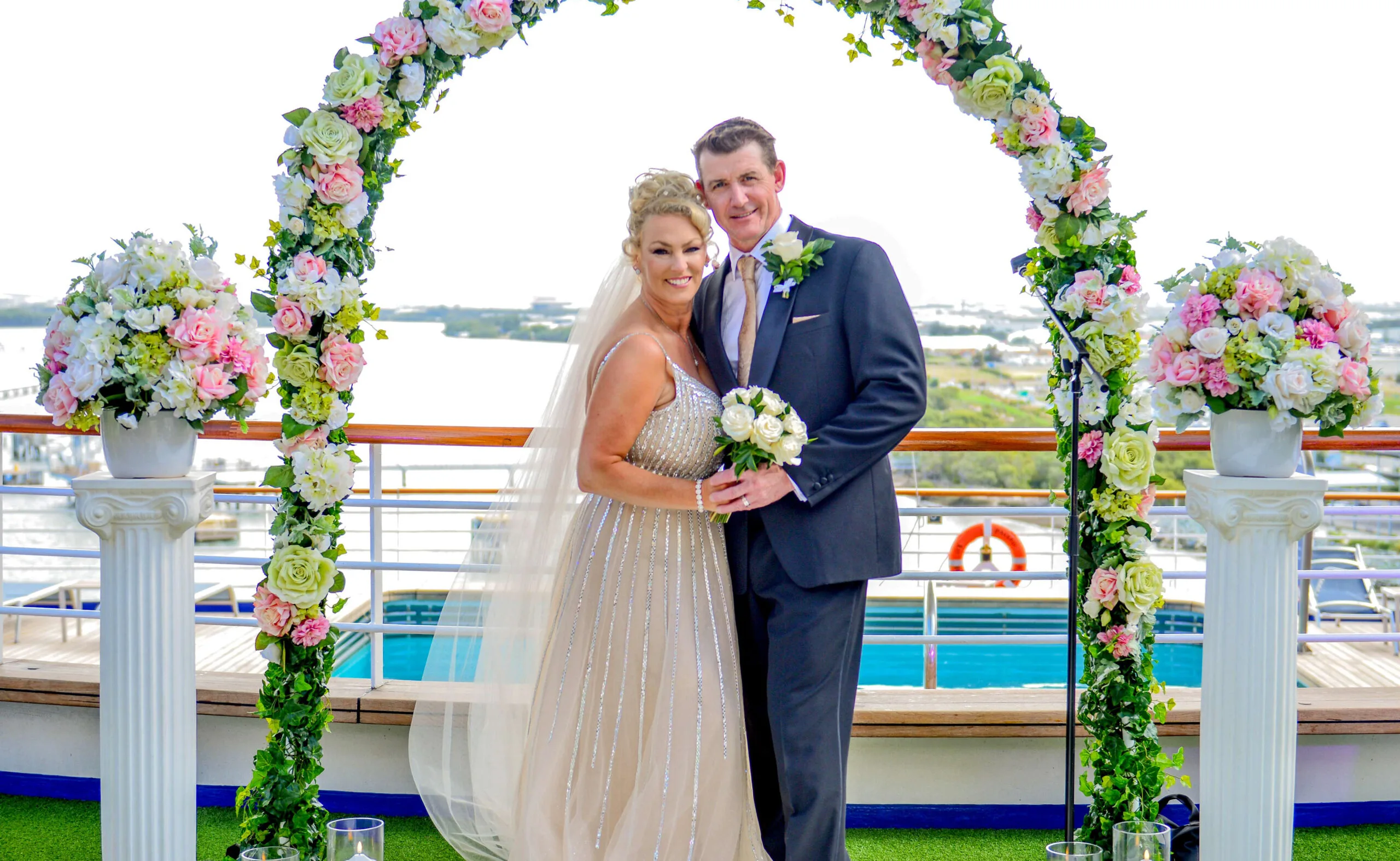 A couple in wedding attire poses under a floral arch on a ship deck with water visible in the background.