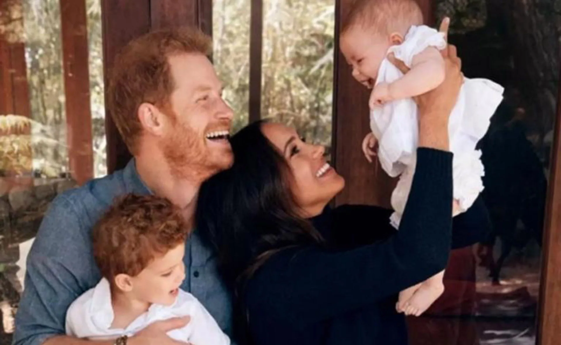 Family smiling, holding two young children, standing in front of a window with trees visible outside.