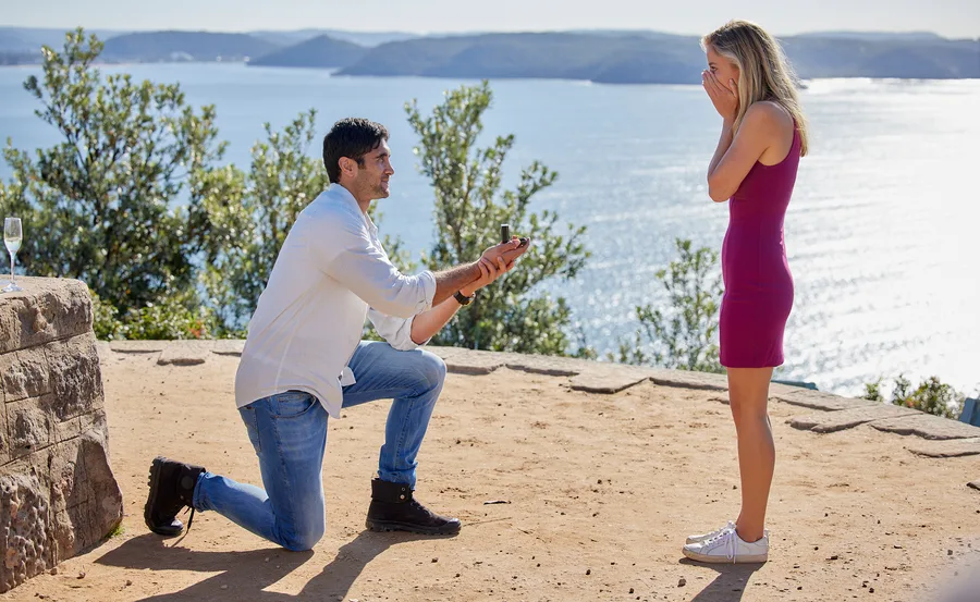 Man kneels holding ring box proposing to surprised woman overlooking sea.
