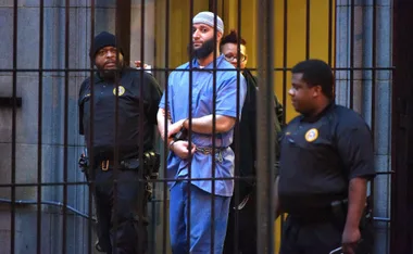 A handcuffed man escorted by three officers outside a building, seen through bars.