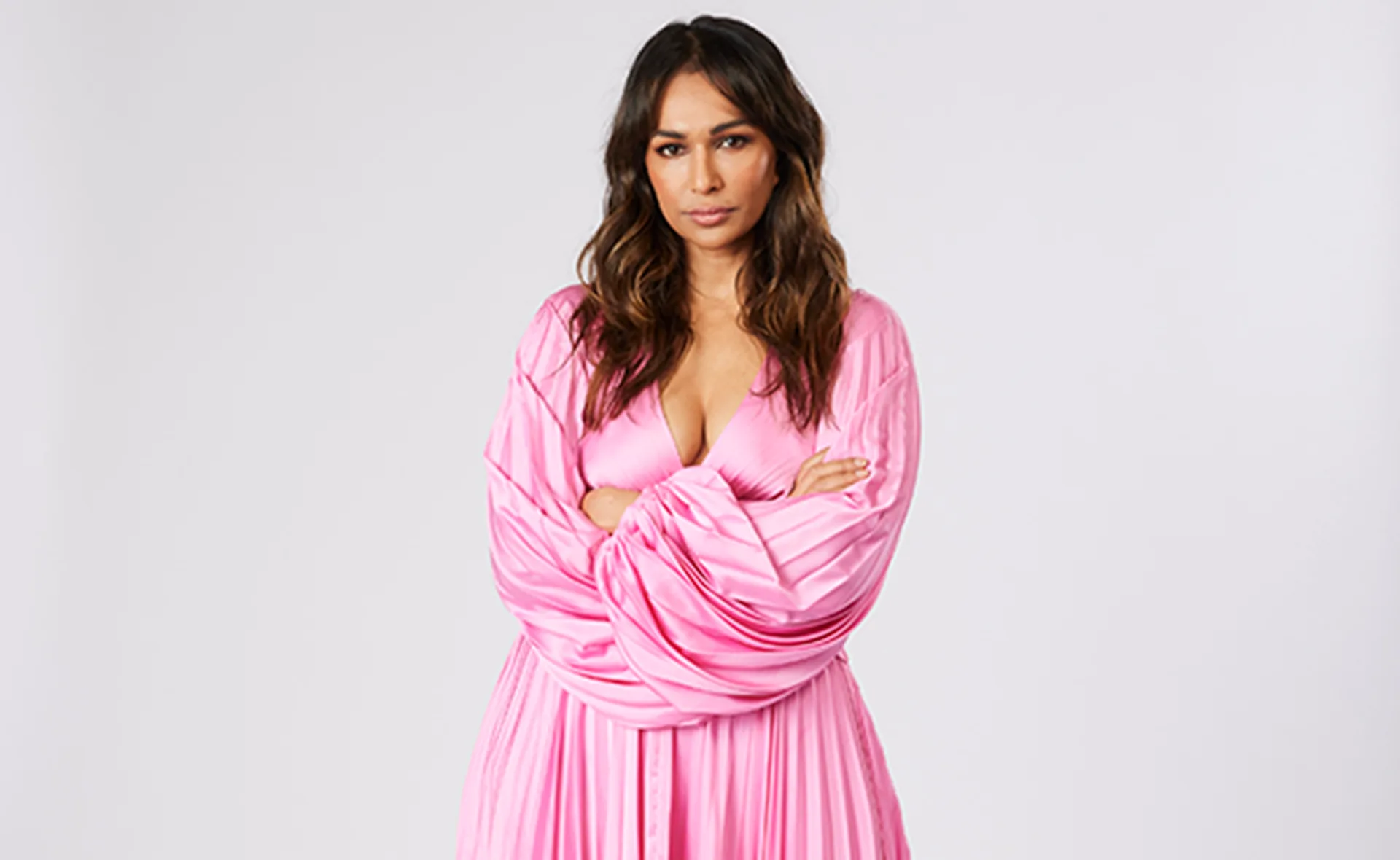Woman in a flowing pink dress with long sleeves, arms crossed, standing against a plain white background.