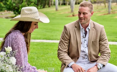A woman in a cowboy hat and man in a beige blazer sit outdoors on grass, smiling, with white flowers in the foreground.