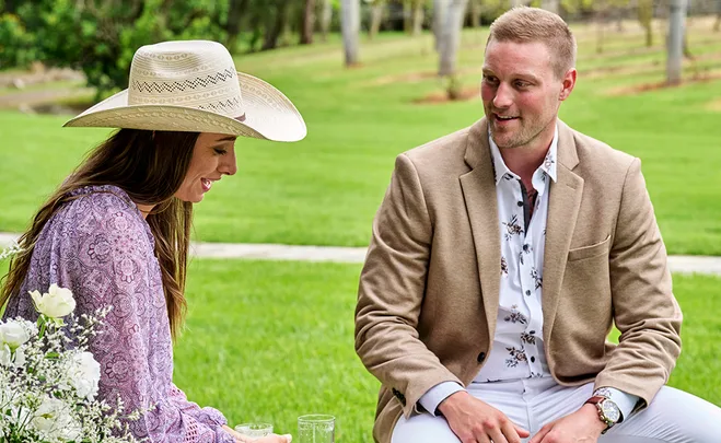 A woman in a cowboy hat and man in a beige blazer sit outdoors on grass, smiling, with white flowers in the foreground.