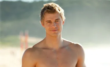 Shirtless man on a sunny beach with a relaxed expression and tousled hair.