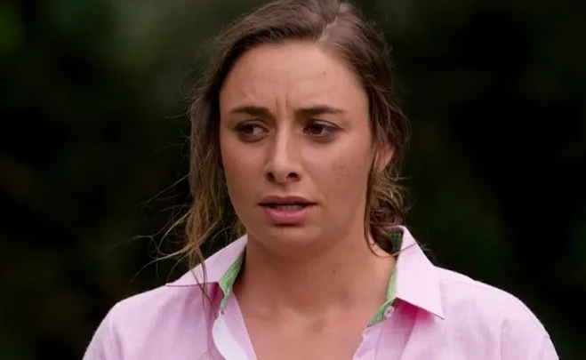 A woman with a concerned expression wearing a pink shirt, standing outdoors with greenery in the background.