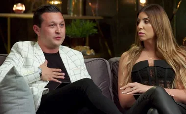 A man in a white jacket gestures while talking, seated next to a woman in a black outfit on a couch indoors.
