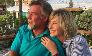 A man and woman smiling and looking into the distance while seated outdoors at a restaurant, surrounded by plants.
