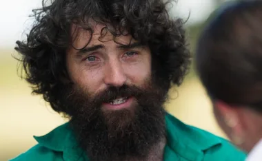 A bearded man with curly hair wearing a green shirt outdoors, talking to another person whose back is visible.