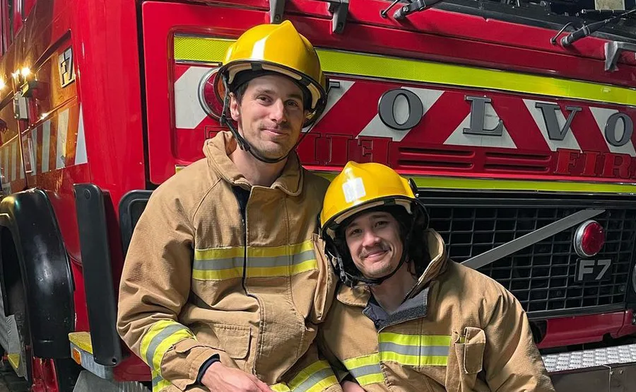 Two firefighters in gear, smiling, sit in front of a red fire truck with "Volvo" visible.