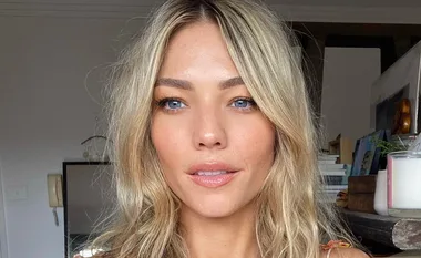 A person with long blonde hair and blue eyes is seen indoors, looking at the camera.