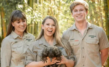 A group of three people wearing Australia Zoo uniforms, one holding an echidna, smiling outdoors.