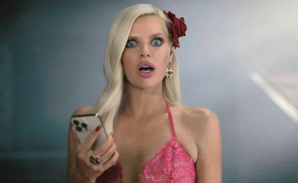 A surprised woman with a red flower in her hair holds a smartphone, wearing a pink lace top.