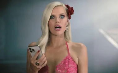 A surprised woman with a red flower in her hair holds a smartphone, wearing a pink lace top.