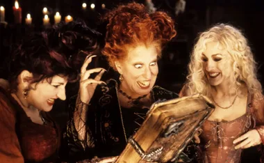 Three witches gathering happily around a magical book with candles flickering in the background.