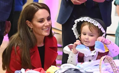 A woman in a red coat smiles at a child holding blue items, wearing a bonnet and costume.