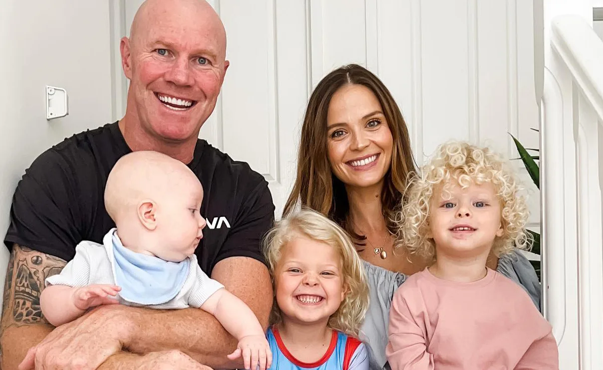 A smiling family group photo with two adults and three young children sitting on stairs indoors.