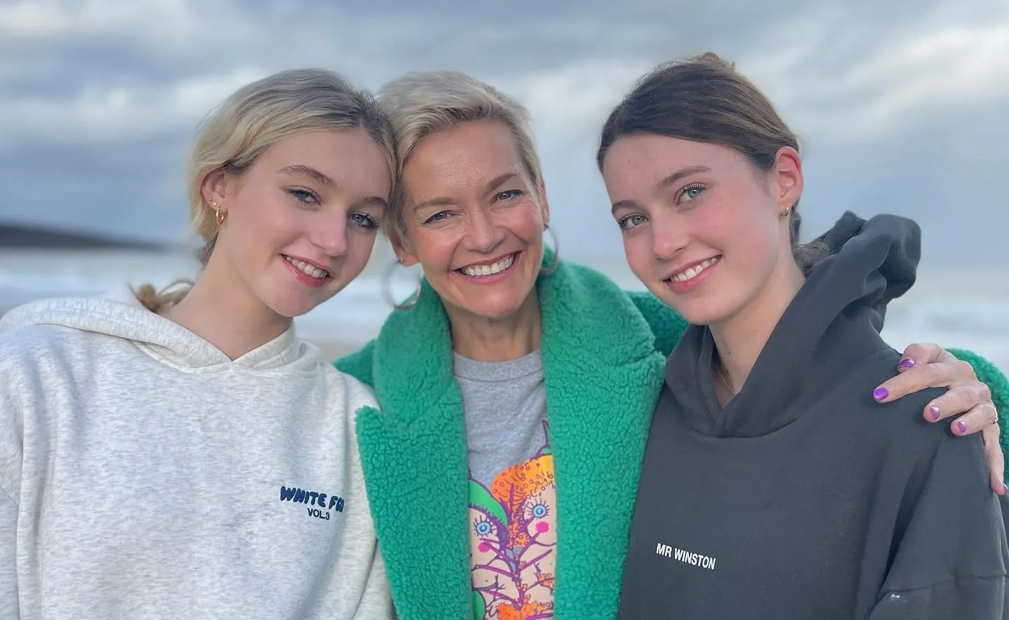 Three individuals smile on a beach, with one wearing a green coat and the others in hoodies.