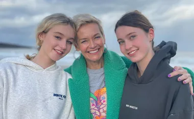 Three individuals smile on a beach, with one wearing a green coat and the others in hoodies.