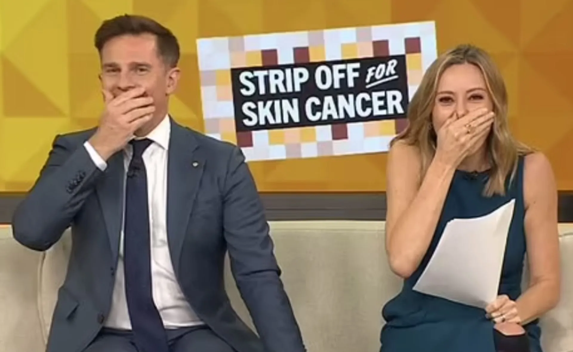 Two people smiling, covering their mouths, sitting in front of a "Strip Off for Skin Cancer" sign on a TV show set.