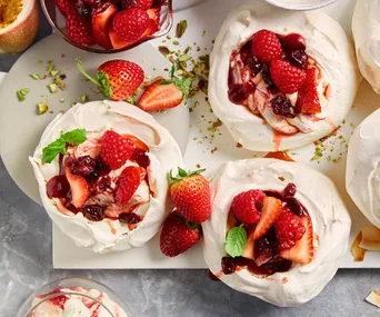 Mini pavlovas topped with raspberries, strawberries, and mint leaves; scattered pistachios and fruits in background.