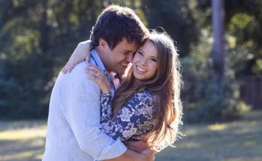 A couple embracing outdoors, smiling, with blurred greenery in the background.