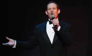 Man in a tuxedo holding a microphone, speaking on stage.