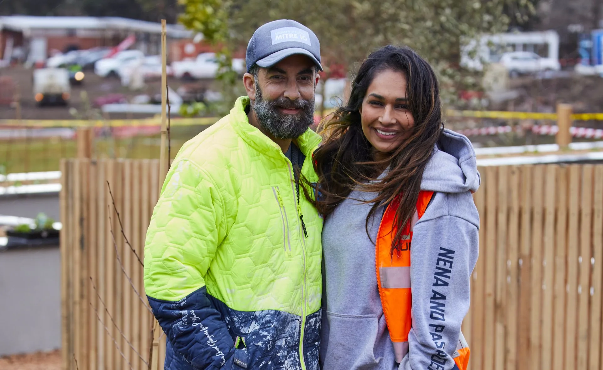 A man in a bright jacket and cap stands smiling with a woman in a hoodie and orange vest, outdoors with a blurred background.