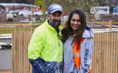 A man in a bright jacket and cap stands smiling with a woman in a hoodie and orange vest, outdoors with a blurred background.