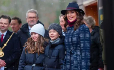 A group of people, including a woman in a checkered coat and hat, are smiling outdoors on a snowy day.