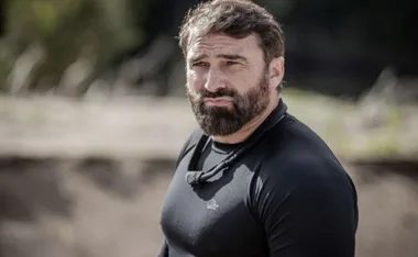 Bearded man in a black shirt outdoors, looking serious.