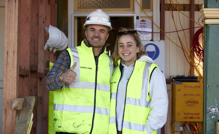 Two people in high-visibility vests and hard hats stand smiling on a construction site, one giving a thumbs-up.