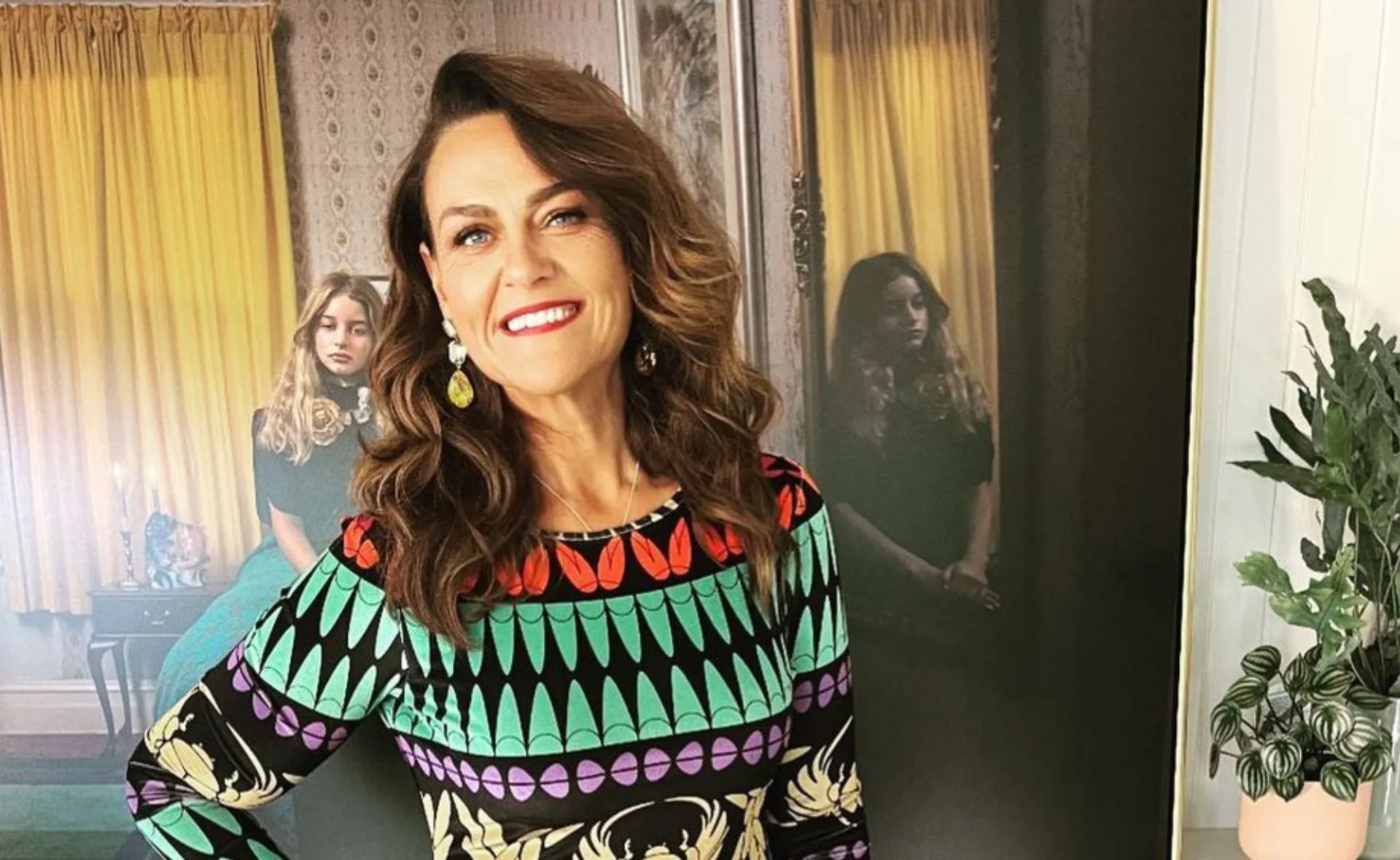 A smiling woman in a colorful dress poses indoors, with a painting and a potted plant in the background.
