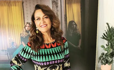 A smiling woman in a colorful dress poses indoors, with a painting and a potted plant in the background.