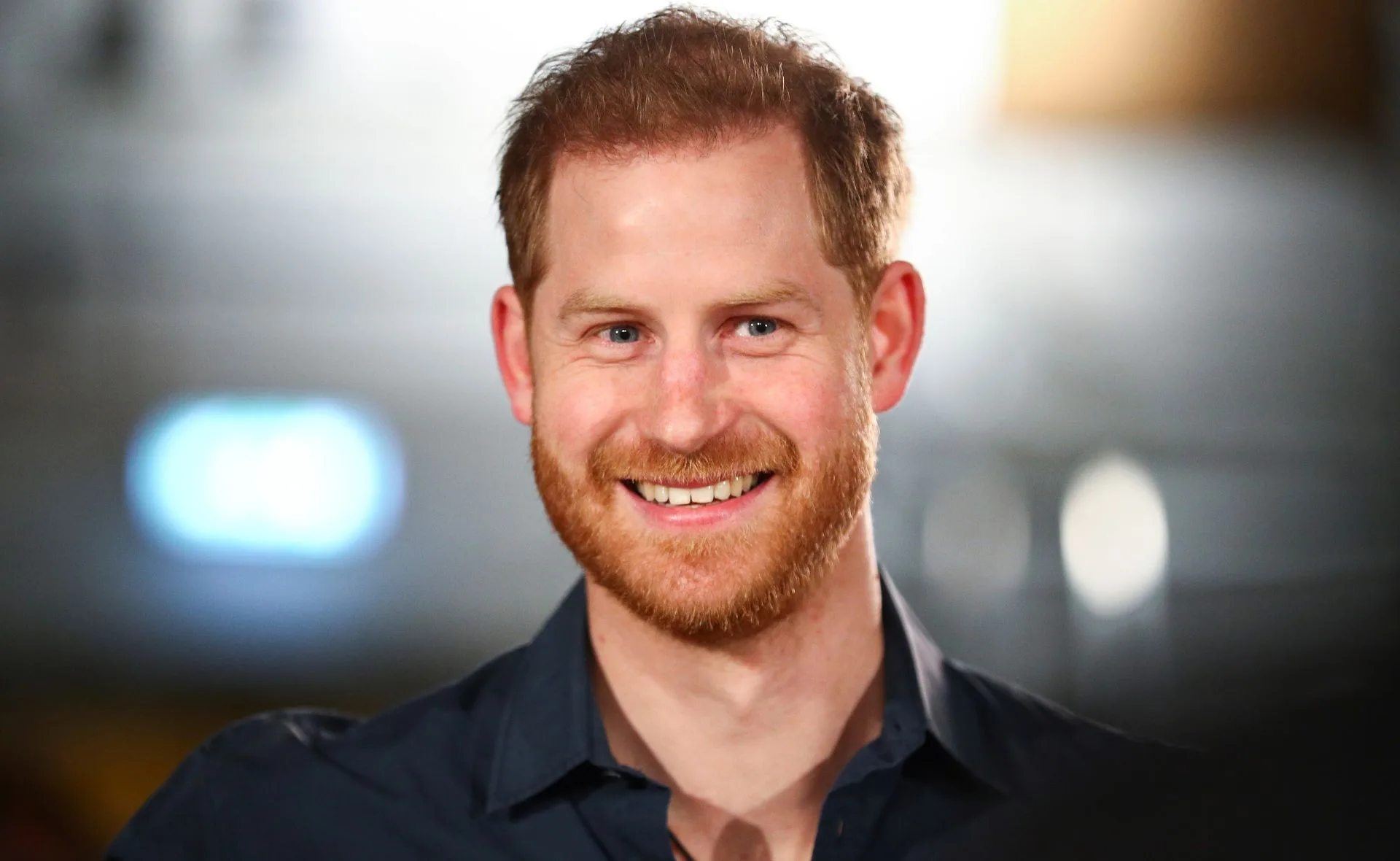 Smiling man with red hair and beard, wearing a dark shirt, with blurred lights in the background.