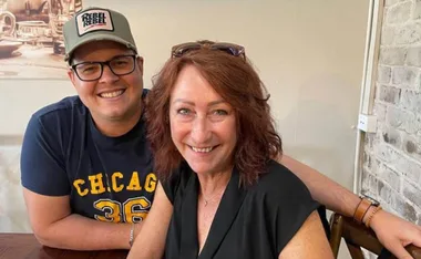 Two people smiling at a table, one wearing glasses and a cap, the other with shoulder-length hair, indoors.