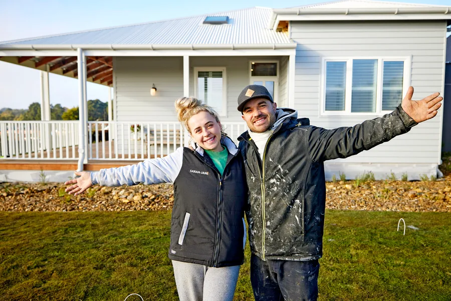 A smiling couple with arms wide open stands in front of a modern house with a porch and white railing.