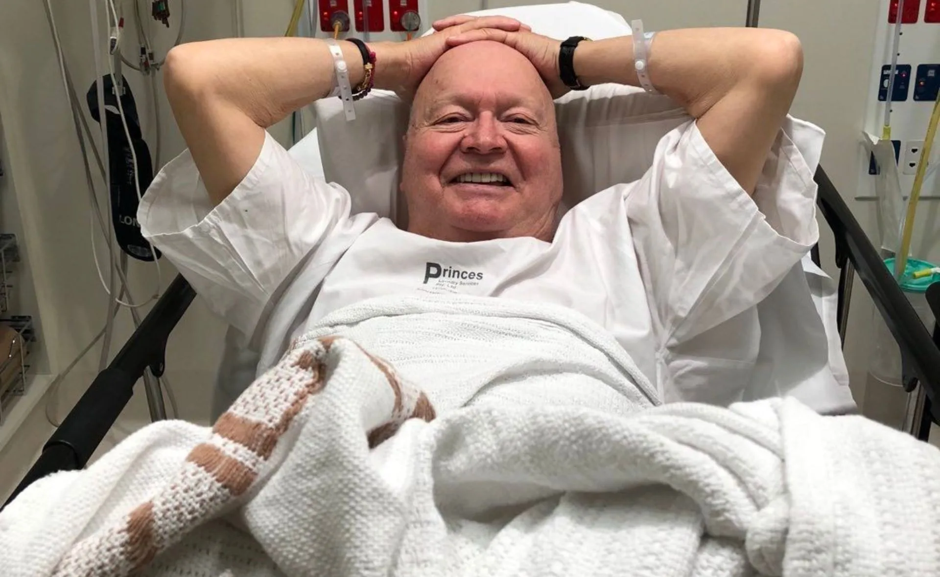 Elderly man smiling, lying in a hospital bed with hands behind his head, wearing a hospital gown.