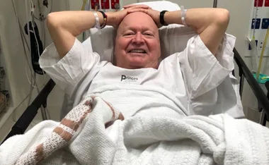Elderly man smiling, lying in a hospital bed with hands behind his head, wearing a hospital gown.