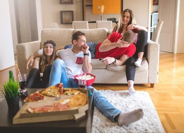 Four friends relaxing on a couch, eating pizza and watching TV in a cozy living room setting.