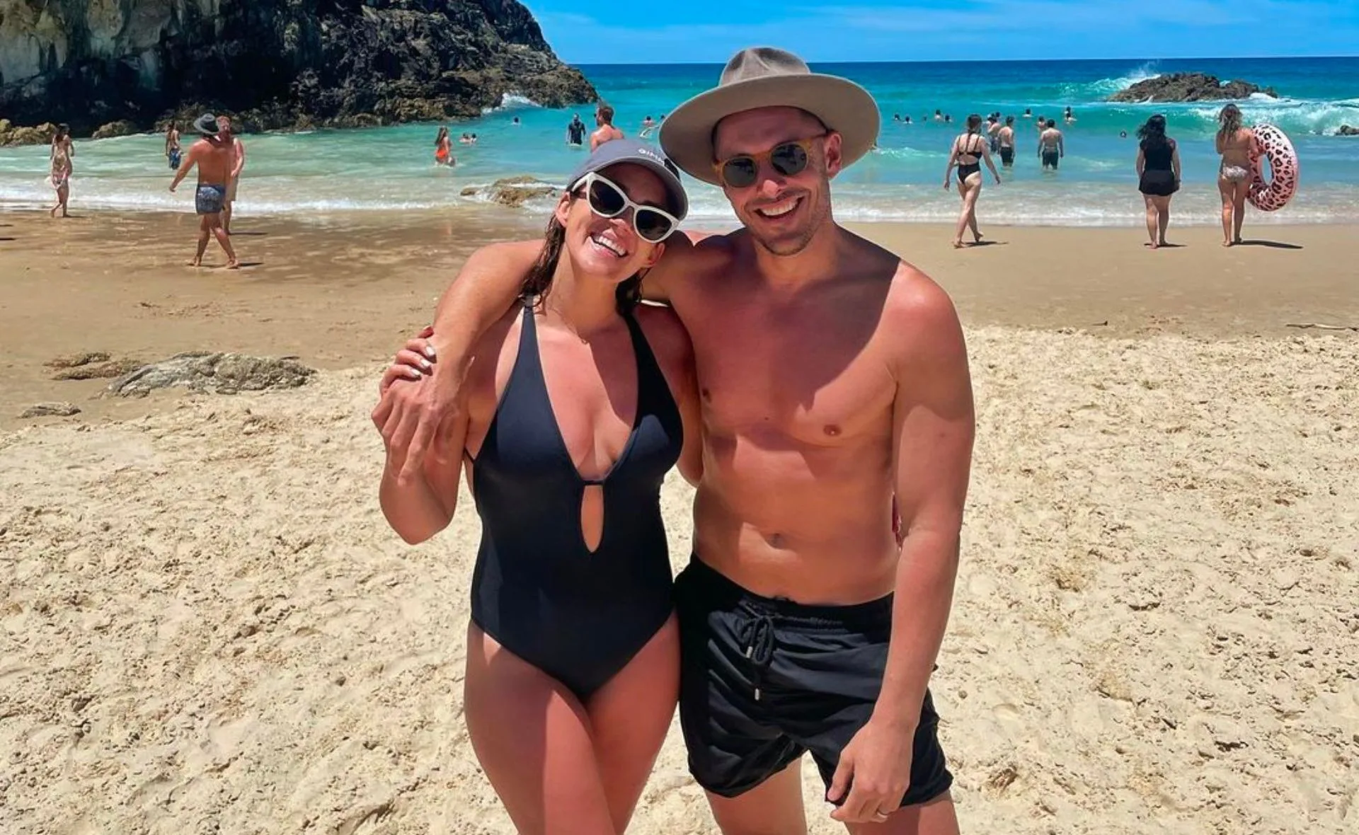 A couple in swimwear smiling at the beach with a blue ocean and clear sky in the background.