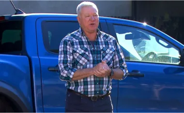 A person in a plaid shirt stands beside a blue vehicle, speaking with hands clasped.