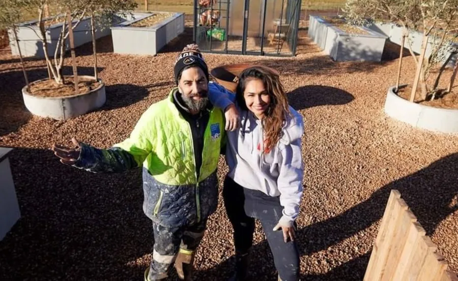Two individuals in casual work attire stand in a garden area with planter boxes and trees.