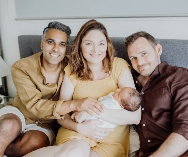 A happy family with two men and a woman holding a newborn baby, sitting on a bed, smiling at the camera.