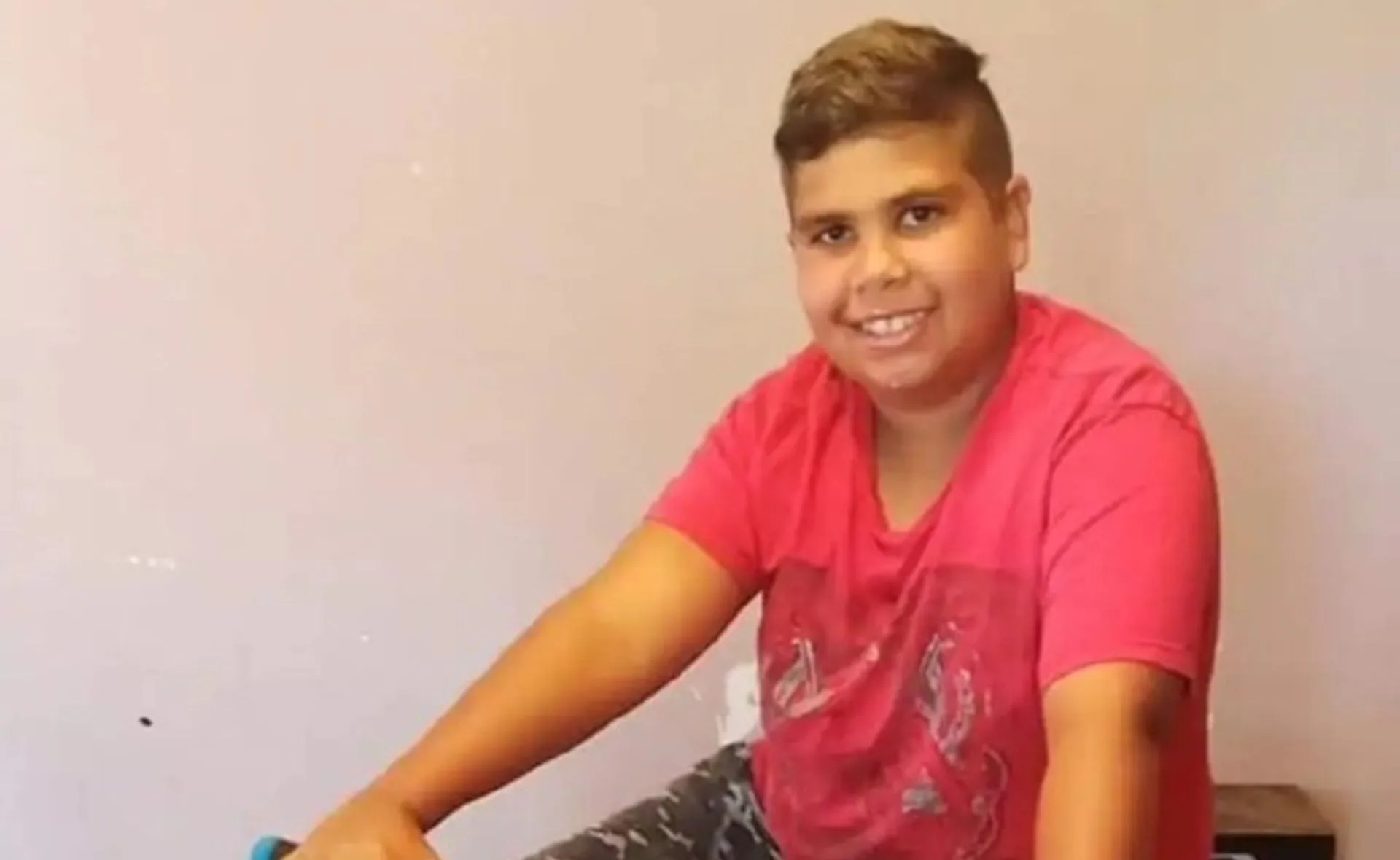 Smiling young boy in a red shirt sits indoors, looking at the camera.
