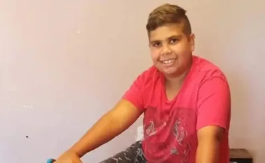 Smiling young boy in a red shirt sits indoors, looking at the camera.