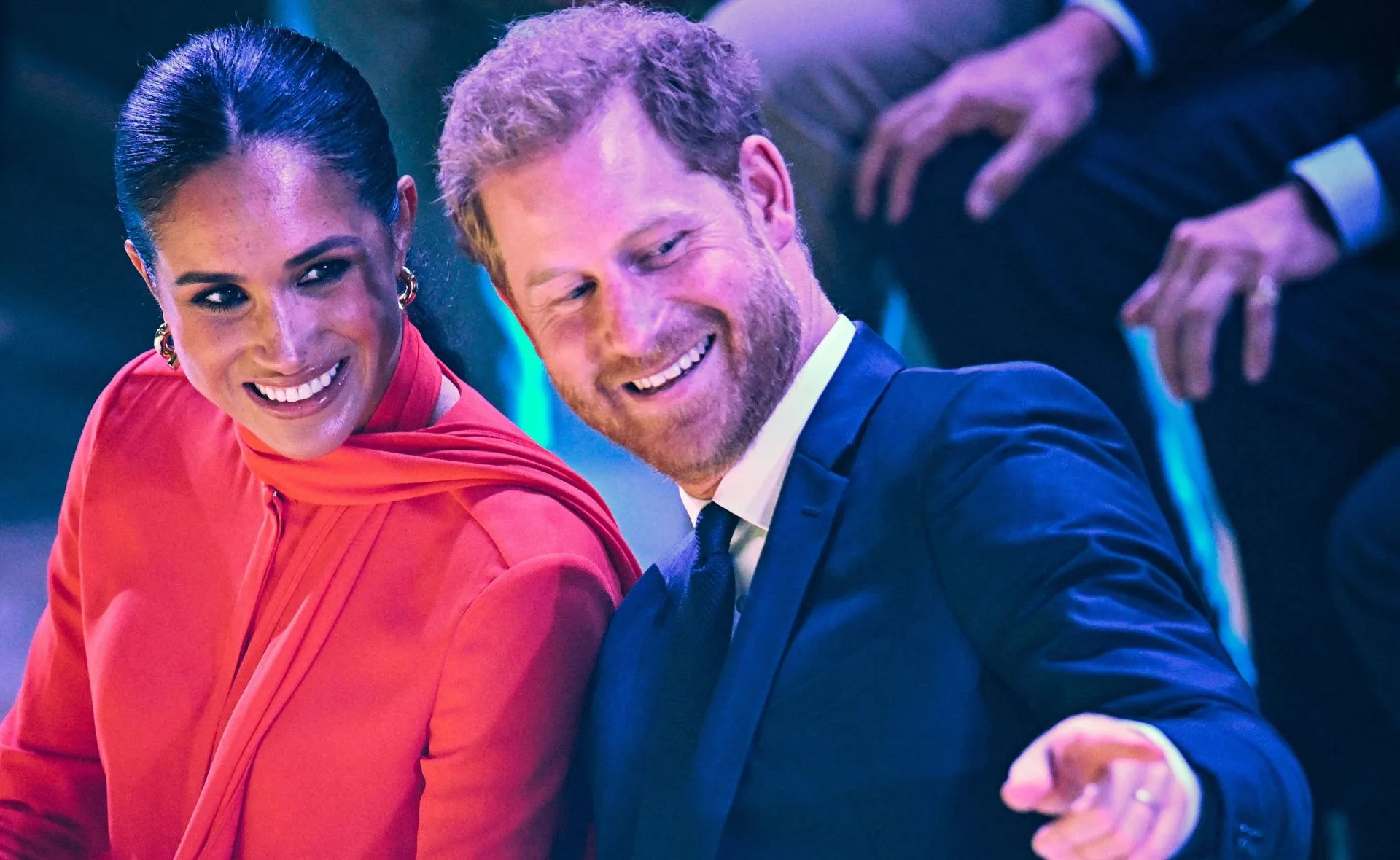 A man in a suit and a woman in a red outfit are sitting together, both smiling and engaged in conversation.