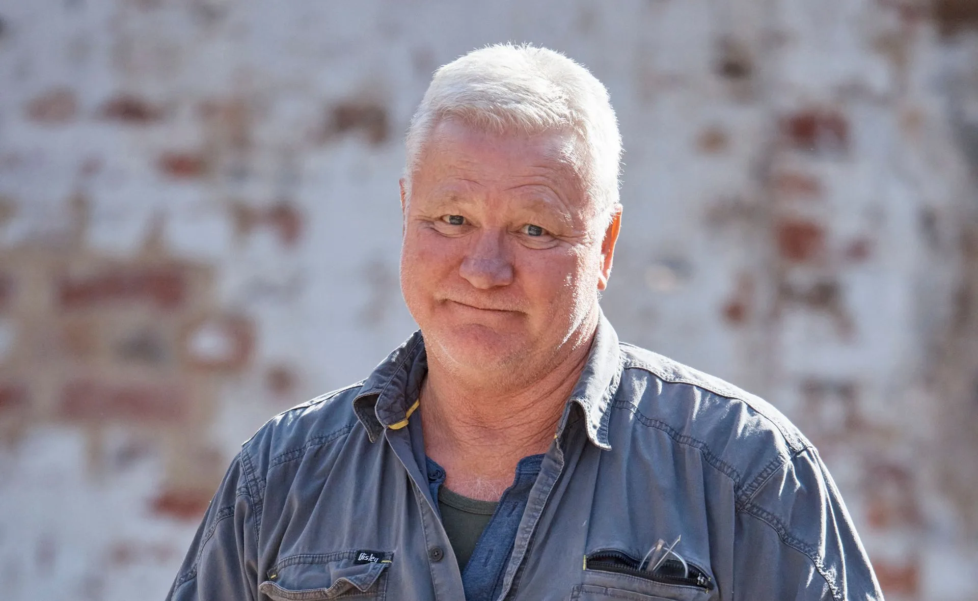 Man with short white hair in a grey shirt stands against a blurred brick background, looking directly at the camera.