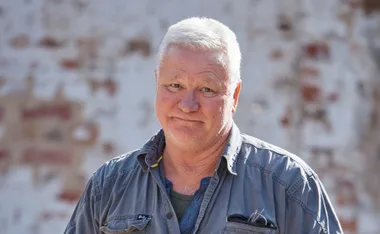 Man with short white hair in a grey shirt stands against a blurred brick background, looking directly at the camera.